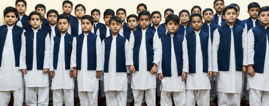 More than thirty boys in white shalwar kameez and navy waistcoats standing together in rows.